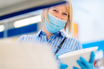 Blond woman wearing disposable medical mask shopping choosing cooking utensils in supermarket,...