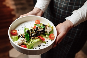 The waiter is holding a white plate with a dish, close-up of hands. Salad with cherry tomatoes, basil and pine nuts