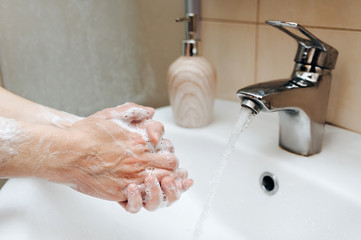 Two female hands with soapy foam in a stream of tap water in the bathroom. The concept of hygiene, cleanliness and protection against coronavirus COVID-19.