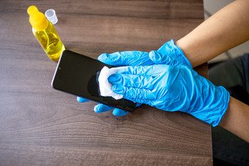 Woman's hand in blue gloves sanitizing cleaning smartphone mobile phone with alcohol on wood table surface with wet wipes