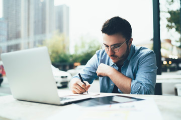 Serious man writing documents sitting at table with laptop in outside cafe