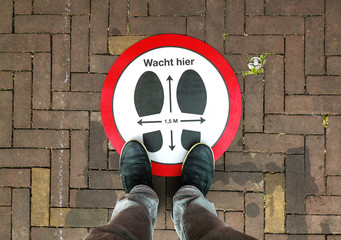A person standing on a social distancing sign. Translation of Dutch text on sign: "Wait here", "1.5m".