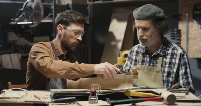 Family of carpenters. Male generations of handicraftsmen. Old father joiner teaching his handsome young son to proceed wood and work with timber. Male craftsmen polishing plank. Planked hardwood.