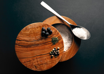 A top view of a wooden bowl of salt with a spoon and some other spices is standing on a black surface