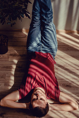 Carefree relaxed man in jeans, wine red t-shirt lying down on parquet floor, feet on wall, hands under head. Natural light and shadow pattern in form of stripes. Stress free concept. Morning sunlight
