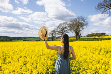 Girl from behind in a field of yellow rapeseed flowers holding a hat in a sunny day