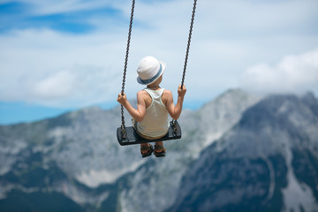 A boy in a summer hat flies on the swings on a background of rocky mountains - obrazy, fototapety, plakaty