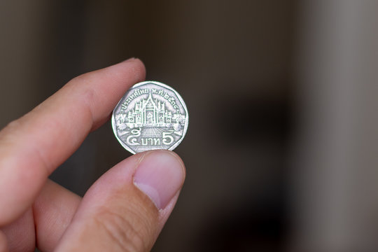 Persons hand holding out a five baht coin, the Currency of the Thailand -  on a brown background. Money exchange.