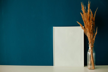 vase with dry grass  and empty white frame on the table on a blue background