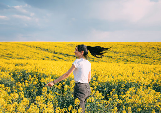 Happy energetic active woman dancing in flowering field. Brunette girl, enjoy beauty spring nature. Hairstyle long black hair gathered in ponytail, fluttering. Backdrop yellow rape flowers, blue sky