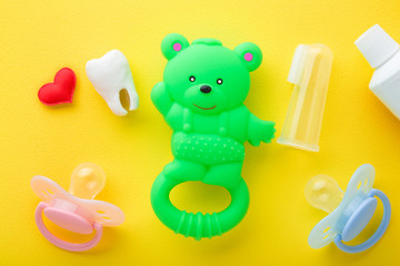 Rubber teddy bear, red heart, tooth, tube of toothpaste, soothers and transparent silicone fingertip toothbrush on yellow table background. Things for babies teeth cleaning through fun. Top down view.