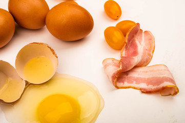 Broken chicken egg, scattered chicken eggs, slices of sausage and bacon and tomatoes on a white background. Close up.