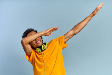 Cheerful African-American young guy joyfully raises hands up covering face as dub dancing sign posing on blue background with headphones in yellow t-shirt. Сoncept of music subscription, online radio