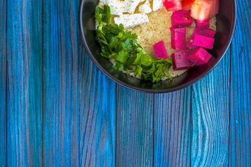 Bowl with porridge of cous cous and vegetables, tomatoes, cheese red and feta, greens, parsley. Useful vegetarian breakfast. Blue wooden background.