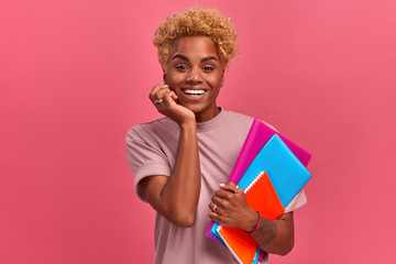 Emotional cheerful young african-american student girl holding notebooks and preparing for entrance exams on pink background. Concept of preparation for study and distance education. Advertising space