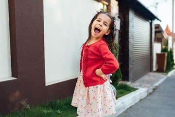 Image of cute little girl laughing during coming home after school day stading near her home outside. Smiling kid playing outdoor, has joyful expression. Happy child smiling broadly and posing outdoor
