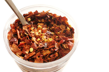 Dried red chili pepper flakes on a plastic jar on white background with copy space on the right side.  Crushed chillies with spoon.  Selective focus.