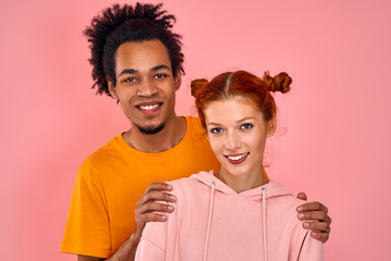 Close-up positive beautiful mixed race couple charming red-haired girl and African American guy posing in studio on pink background. Concept of love relationships, multicultural. Advertising space