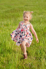 A happy little girl in summer dress with pigtails run in a green field, light of warm rays of the sun