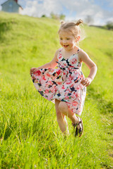 A happy little girl in summer dress with pigtails run in a green field, light of warm rays of the sun