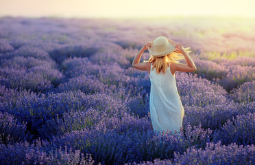 Beautiful sunset on a lavender field. Romantic lady wears a straw hat, sunrays are shining on her face. Beautifully fluttering hair, warm summer day. Freedom and psychological comfort concept