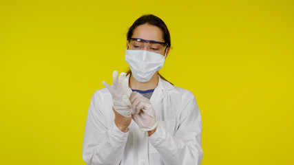 Young doctor in a medical mask and safety glasses puts latex gloves on her hands. Girl in a white coat on yellow background