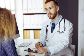 Friendly red-bearded doctor and patient woman discussing current health examination while sitting...