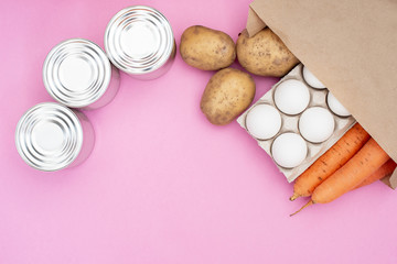 Food donation for the needy on a pink background top view. Potatoes, carrots, water, milk, oil, eggs and canned food.