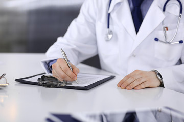 Unknown male doctor sitting and working with clipboard of medication history record in clinic at his working place, close-up. Young physician at work. Perfect medical service, medicine concept