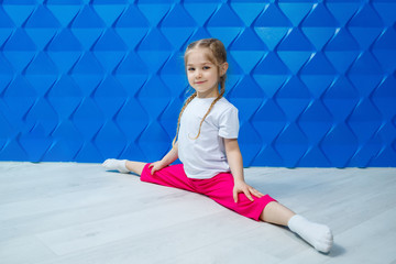 A little girl with pigtails in a white T-shirt on a blue background sits in twine on the floor and smiles. Children's emotions occupation spotrom, fun