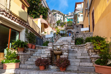 Colorful narrow stairs street with Fountain and flowers near square Piazza Duomo in Taormina in sunny morning, Sicily, Italy
