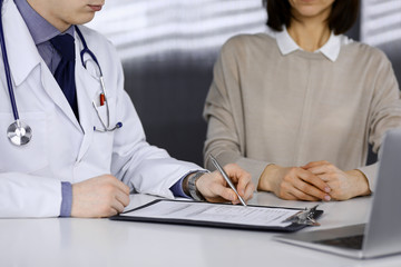 Obraz premium Unknown male doctor and patient woman discussing something while sitting in clinic and using clipboard. Best medical service in hospital, medicine, pandemic stop