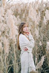 portrait of a young beautiful girl in a white dress, a warm knitted cardigan and black shoes among dry fluffy reeds in the autumn time at sunset.nature,fashion concept