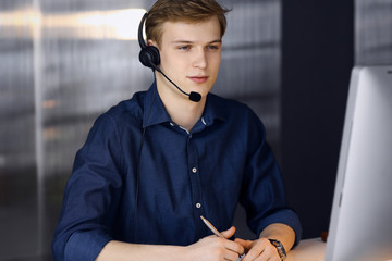 Young blond businessman using headset and computer at work. Startup business means working hard and...