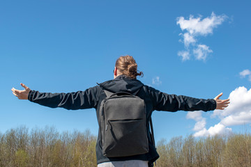 A man from the back with arms outstretched against the sky. A man with long hair and a backpack on his shoulders in nature feels free.