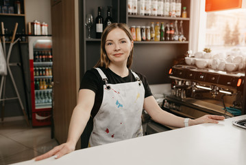 A female barista with a white apron poses behind the bar in a cafe. A pretty female business owner in her coffee shop.