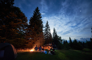 Fototapeta premium Group of travelers sitting near bonfire in forest with tall coniferous trees, car and camp tent under beautiful night sky. Fantastic view of blue starry sky over meadow with travelers near campfire.