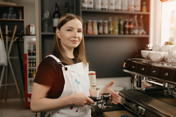 A female barista with a white apron poses holding a metal tamper and a portafilter with coffee in a coffee shop. A pretty female business owner near a professional espresso machine in her cafe.