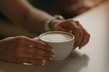 A girl with beautiful nails keeps a cup of latte in her hands in a cafe. 