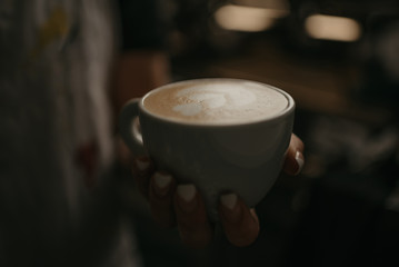 A female barista holds a cup of hot latte in her hand in a cafe. A waitress preparing a customer order in the evening.