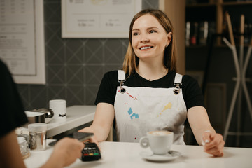 A close portrait of a female barista who smiling holds out to a client a terminal for paying. A woman paying for a cup of latte with a smartphone by contactless PAY PASS technology in a cafe. 