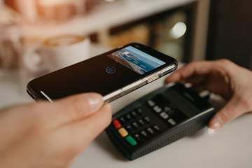 A girl paying for her latte with a smartphone by contactless PAY PASS technology in a cafe. A female barista holds out a terminal for paying to a client in a coffee shop.