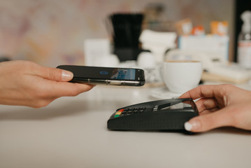 A woman preparing to pay for her latte with a smartphone by contactless NFC technology in a cafe. A female barista holds out a terminal for paying to a client in a coffee shop.