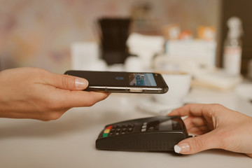 A close-up photo of a smartphone preparing to pay for a latte by contactless PAY PASS technology in a cafe. A female barista holds out a terminal for paying to a client in a coffee shop.