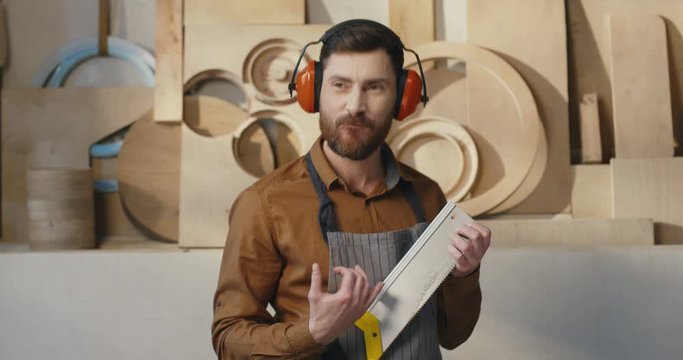 Cheerful young handsome caucasian male carpenter in headphones having fun and playing on saw like on the guitar. Joyful man handicraftsman holding file and play music.