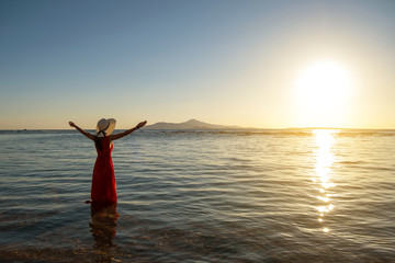 Young woman wearing long red dress and straw hat raising hands standing in sea water at the beach...