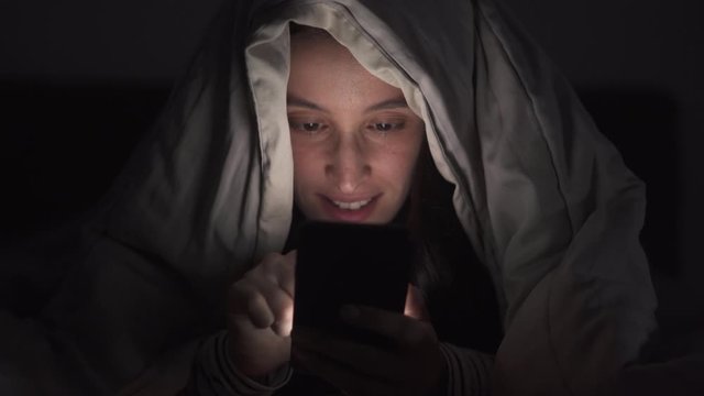 Woman is browsing social media in mobile phone at night under the blanket, Face close-up.
