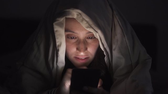 Woman is browsing social media in mobile phone at night under the blanket, Face close-up.
