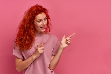 Naklejka premium Joyful beautiful red-haired young girl student points finger to left on pink background in the studio. Concept of advertising household appliances in an online store. Advertising space