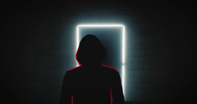 Unrecognizable Silhouette Of A Person With A Hood Stands In Front Of A Glowing Frame Of A Door In The Dark Corridor. Zoom In. Digital Animated.